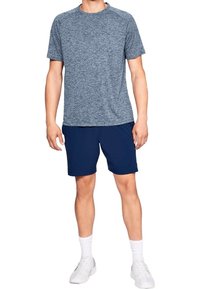 Hombre de pie con una camiseta de manga corta gris jaspeado, pantalones cortos azul marino, calcetas blancas y zapatillas blancas, sobre un fondo blanco.