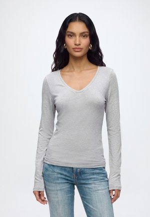 Femme aux longs cheveux foncés portant des boucles d'oreilles créoles dorées, un t-shirt gris clair à manches longues avec col en V, et un jean bleu délavé, debout devant un fond blanc.