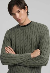 Maglione verde oliva a trecce con scollo rotondo, caratterizzato da motivi trecciati in rilievo e polsini a coste. Indossato sopra una camicia nera.