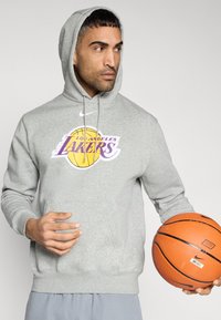 Sweat à capuche gris avec une poche avant, arborant un logo des Los Angeles Lakers en violet et jaune. Comprend une capuche avec cordon. Tenant un ballon de basket.