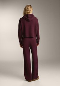 Žena s dlhými blond vlasmi, oblečená v tmavo fialovej mikine s kapucňou a zodpovedajúcich širokých nohaviciach, stojí s chrbtom k fotografii.