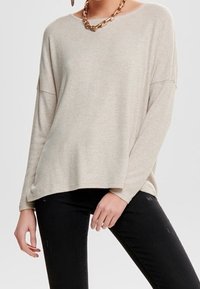 Pull en tricot beige à coupe décontractée, avec des manches longues et un col large. Porté avec un jean foncé et un collier chaîne épais.