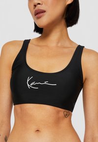 Scollo ampio e bretelle larghe, reggiseno sportivo nero realizzato in materiale elasticizzato, con un design firmato bianco sul davanti.