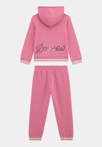 Survêtement rose en deux pièces comprenant un sweat à capuche avec un logo "Guess" pailleté et un pantalon assorti avec des accents rayés aux poignets.