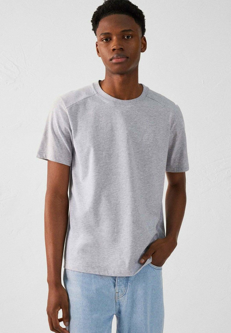 Camiseta gris de manga corta hecha de tejido suave, con un cuello redondo y una forma ajustada. Textura suave con un detalle de marca sutil.