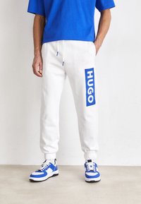 Weiße Jogginghosen mit blauem Logo an der Seite, kombiniert mit einem blauen Shirt und blauen und weißen Sneakern mit schwarzen Akzenten.