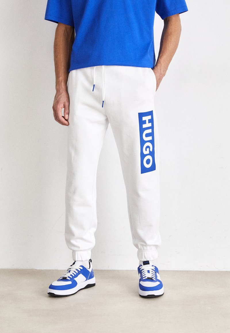 Weiße Jogginghosen mit blauem Logo an der Seite, kombiniert mit einem blauen Shirt und blauen und weißen Sneakern mit schwarzen Akzenten.