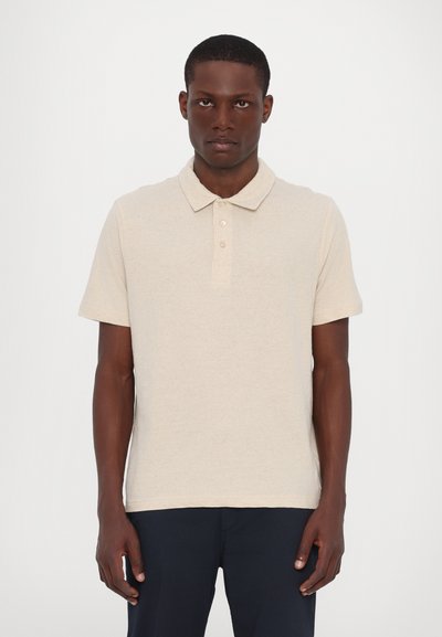 Beige Poloshirt mit kurzen Ärmeln, einer Knopfleiste mit drei Knöpfen und einer weichen Textur. Getragen mit dunklen Hosen, stehend vor einem neutralen Hintergrund.