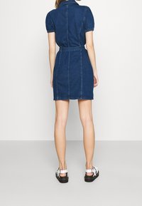 Kvinna som står med ryggen mot, iförd en kort mörkblå denimklänning med puffärmar och vita sandaler med svarta sulor.