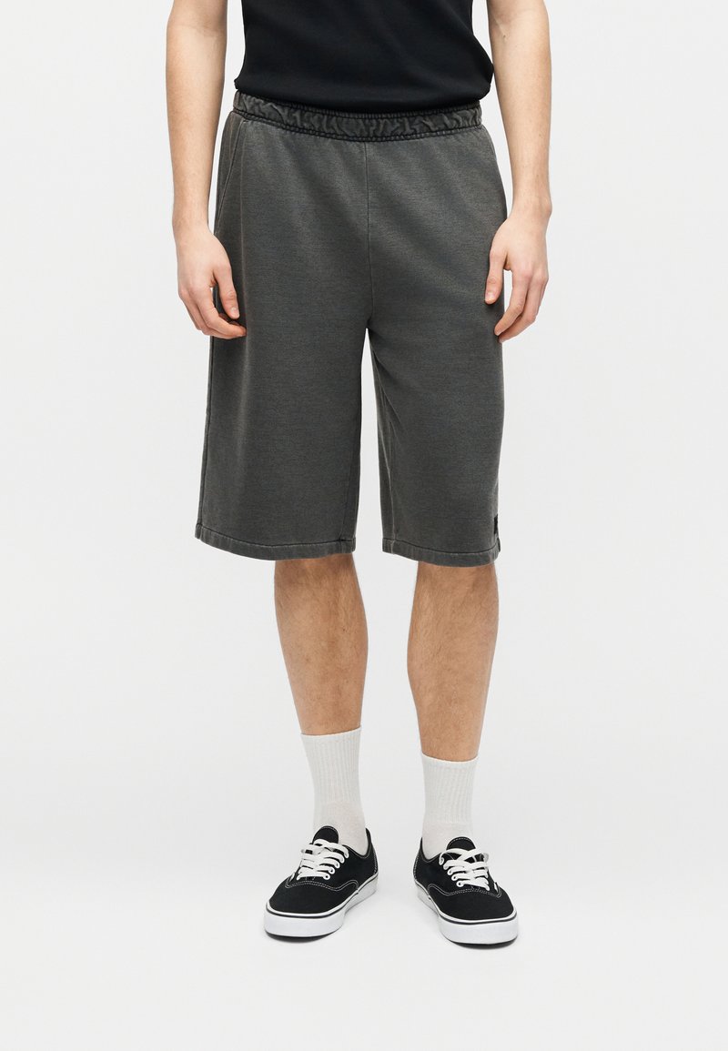 Persona con pantalones cortos gris oscuro hasta la rodilla, zapatillas negras con cordones blancos, calcetines blancos y una camiseta negra sobre un fondo liso.