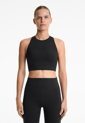 Donna con pelle chiara e occhi azzurri, che indossa un top corto senza maniche nero e leggings neri a vita alta, in piedi di fronte a uno sfondo bianco.