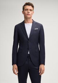 Marineblauer Anzug mit zwei Knöpfen, Reverskragen und aufgesetzten Taschen; kombiniert mit einem weißen T-Shirt; glatte Stoffstruktur und taillierte Passform.