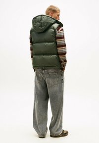 Dunkelgrüne Steppweste mit Kapuze, getragen über einem gestreiften Langarmshirt. Kombiniert mit lockeren, hellgrauen Jeans und braunem Schuhwerk.