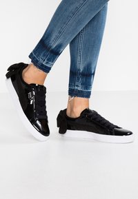 Sneaker neri con finitura lucida in vernice, design con lacci, dettagli in camoscio e un fiocco sul retro. Suola in gomma bianca.