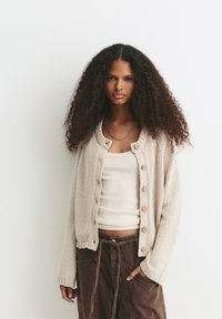Jeune femme avec des cheveux bouclés volumineux portant un cardigan crème, un débardeur beige et un pantalon marron avec une ceinture tressée, se tenant contre un mur blanc.
