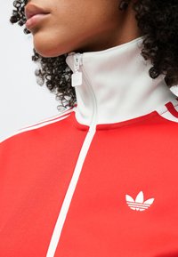 Tuvplānā persona, kas valkā sarkanu Adidas jaku ar baltu augstu apkakli un rāvējslēdzēju, redzams Adidas logotips uz krūtīm.