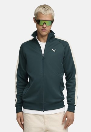 Puma Zip-Jacke in Dunkelblau mit cremefarbenen Seitenpanelen und Logo. Verfügt über zwei Fronttaschen und einen hohen Kragen. Trägt man über einem weißen Shirt.