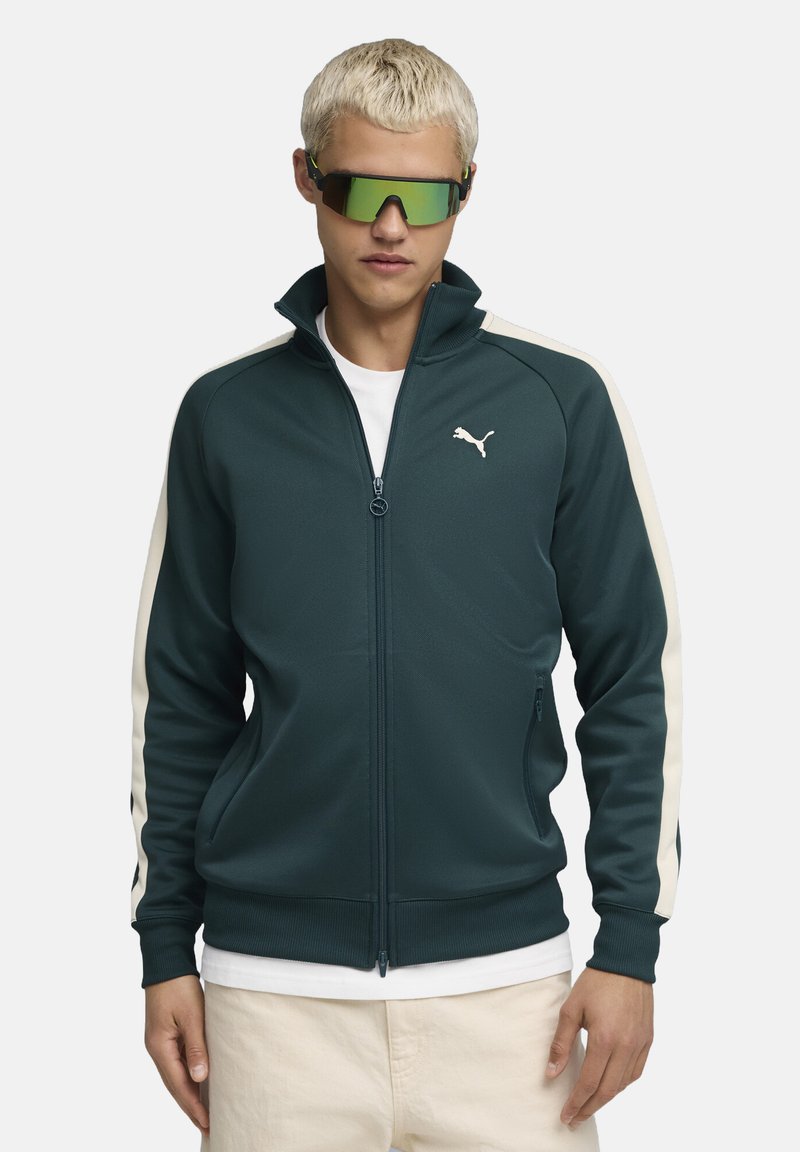 Puma Zip-Jacke in Dunkelblau mit cremefarbenen Seitenpanelen und Logo. Verfügt über zwei Fronttaschen und einen hohen Kragen. Trägt man über einem weißen Shirt.