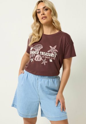 Kvinde med langt blond hår iført en vinrød "Summer Treasures" t-shirt med muslinger og søstjerner og lyseblå denimshorts.