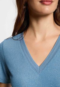 Top blu con scollatura a V e maniche corte, realizzato in un tessuto testurizzato con sottili fili metallici, evidenziando un design aderente.