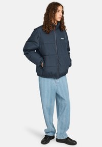 Giovane con lunghi capelli ricci, indossa un giubbotto imbottito blu navy, jeans larghi a fantasia blu chiaro e sneakers nere, in piedi di lato.