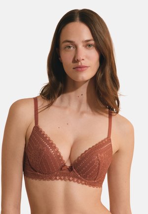 Jeune femme aux longs cheveux bruns portant un soutien-gorge en dentelle couleur rouille, faisant face à un fond clair uni.