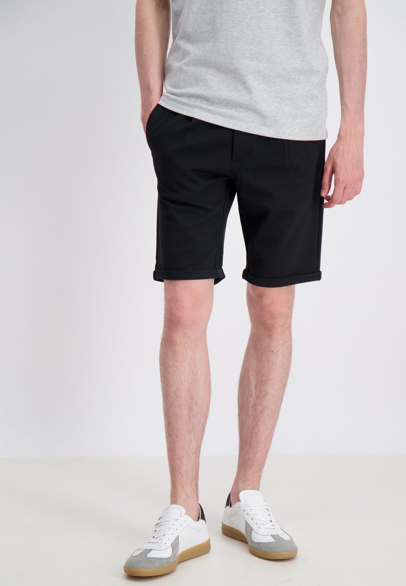 Pantaloni shorts neri realizzati in un tessuto liscio, con orli arrotolati e tasche. Abbinati a sneakers bianche e grigie.