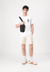 Balta marškinėliai su logotipu, kreminės šortai su kišenėmis, juodas kryžminis krepšys, tan spalvos sportbačiai su raudonomis juostelėmis, stovintis ant vienspalvio fono.