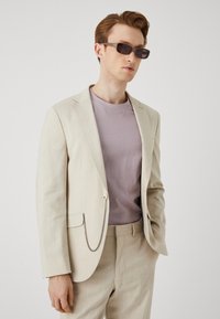 Lichtbeige colbert met een textuurfinish, slim fit, kettingdetail op de zak, gedragen over een lila t-shirt. Donkere zonnebril maakt de look compleet.