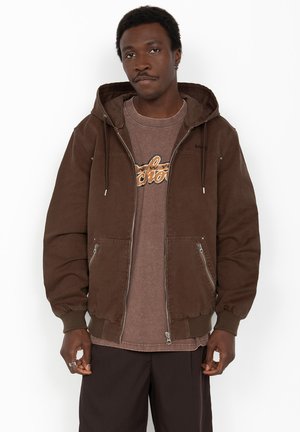 Homme portant une veste à capuche marron avec fermeture éclair sur un t-shirt graphique marron et un pantalon foncé, debout devant un fond blanc.