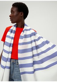 Écharpe rayée en tissu léger, avec des rayures bleues et blanches, drapée sur les épaules, contrastant avec un haut rouge et un jean.