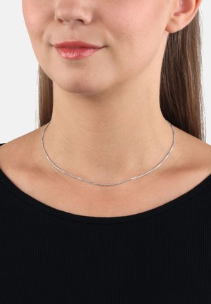 Donna che indossa una sottile collana d'argento su un top nero a coste, che mostra il collo, la parte bassa del viso e i lunghi capelli castani.