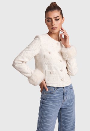 Frau trägt eine weiße, strukturierte Jacke mit Pelzmanschetten und Perlenknöpfen, kombiniert mit hoch taillierten blauen Polka-Dot-Jeans, posiert mit einer Hand im Gesicht.