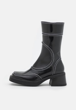 Schwarzer Lederstiefel bis zur Mitte der Wade mit weißen Nähten, quadratischer Kappe und klobigem Blockabsatz, von der Außenseite auf hellem Hintergrund dargestellt.