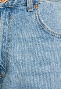 Primo piano di jeans in denim azzurro chiaro che mostra la tasca anteriore, il passante per cintura, i rivetti di rame e il bottone in metallo con dettagli di cucitura.