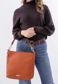 Orange læder tote taske med struktureret overflade, rektangulær form og håndtag i guldfarvet kæde, parret med et brunt bælte og jeans.