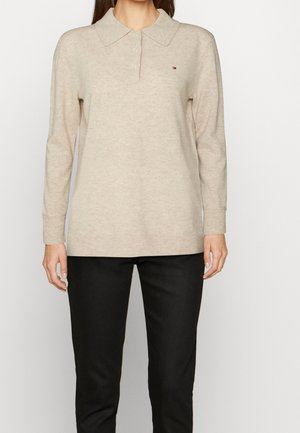 Femme portant un pull beige à manches longues avec col et boutons et un pantalon noir, debout devant un fond uni clair.