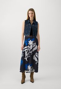 Colete de denim com um design sem mangas, combinado com uma saia longa com grandes padrões florais em azul, cinza e branco sobre um fundo escuro.