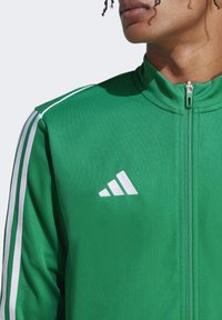 Groene sportjack met witte strepen op de schouders en ritssluiting. Heeft een geborduurd wit logo op de linkerkant van de borst. Soepele textuur.