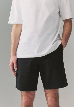 Man draagt een wit shirt met korte mouwen en zwarte korte broek, met één hand in zijn zak en de andere ontspannen langs zijn zijde.