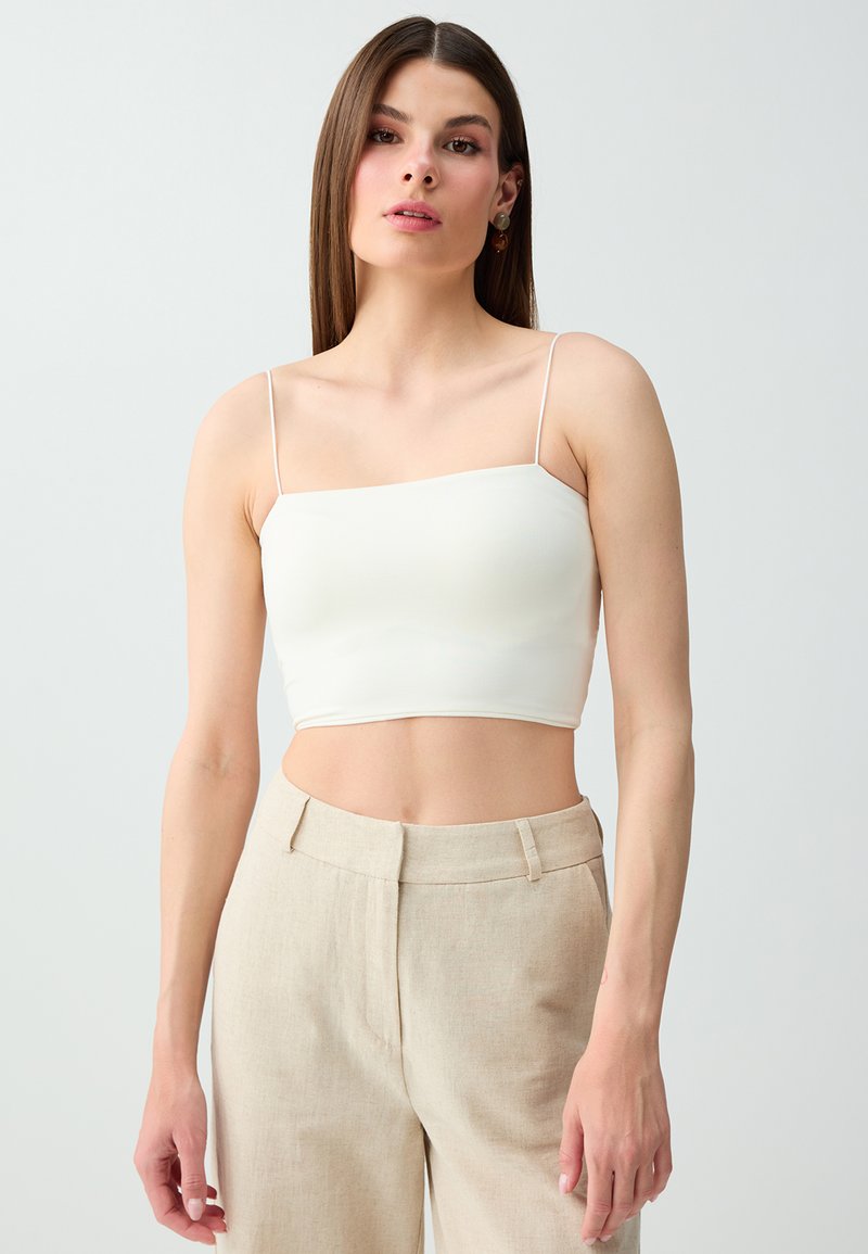 Femme aux cheveux bruns raides portant un caraco court blanc et un pantalon beige taille haute, debout devant un fond clair uni.