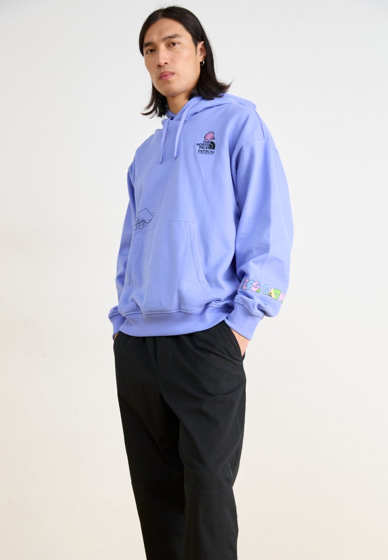 Light purple hoodie with front pocket, featuring a black logo and graphic. Long sleeves with colorful text on the left cuff. Worn with black pants.
