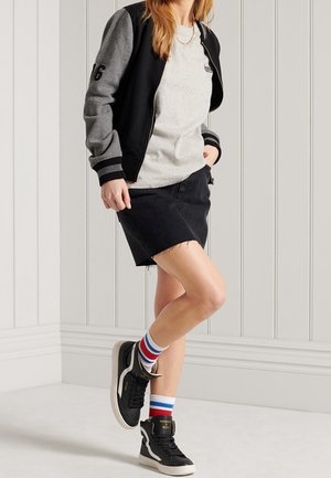 Veste bomber noire avec des manches grises, t-shirt gris, jupe en denim noir et baskets noires avec des chaussettes de cheville rayées rouge, blanc et bleu.