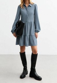 Blå knappad klänning med långa ärmar, lagerdesign och textilierad tyg. Matchas med knähöga svarta stövlar och en svart handväska.