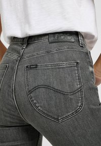 Jean en denim gris avec une finition texturée, comportant une poche arrière avec une petite étiquette noire et des détails de couture subtils.