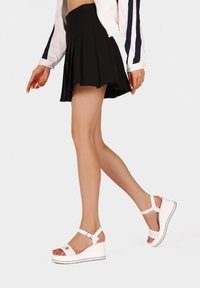 Sandali platform bianchi con cinturino alla caviglia regolabile, suola testurizzata e finitura liscia. Abbinati a una gonna nera plissettata.