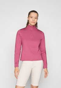 Maglia a maniche lunghe, rosa, con collo zip realizzata in tessuto elastico, caratterizzata da un design aderente e cuciture piatte. Indossata con pantaloni corti di colore chiaro.