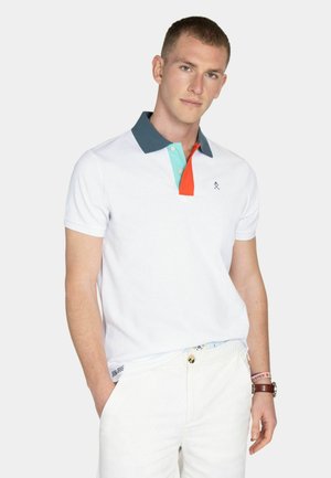 Joven de pie con las manos en los bolsillos, vistiendo una camiseta polo blanca con cuello oscuro y tapeta colorida, combinada con pantalones blancos.
