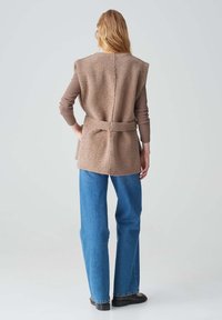 Braun strukturierte Weste mit einem Taillengürtel, geschichtet über einem eng anliegenden Langarmhemd, kombiniert mit blauen, weiten Jeans und dunklen Schuhen.