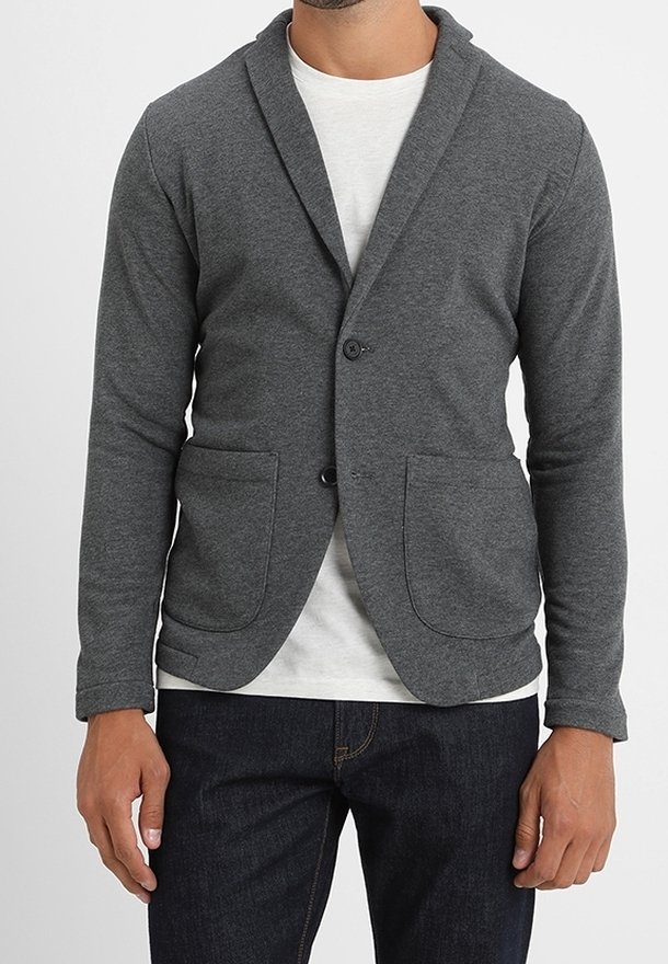Homme portant un blazer gris foncé décontracté sur une chemise blanche et un jean foncé, debout les bras détendus le long du corps.